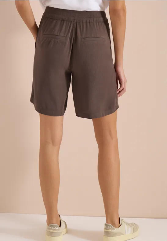 Achteraanzicht van een persoon met casual bruine korte broek met elastische taille en zakken tegen een beige achtergrond.