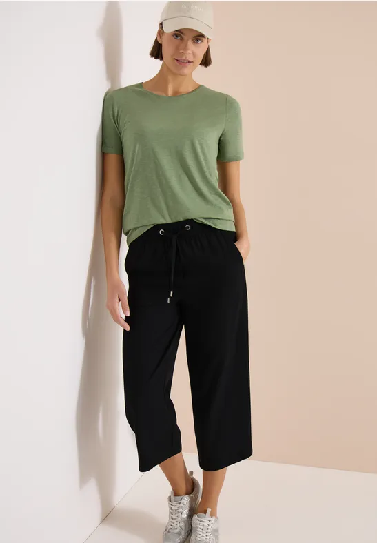 Vrouw in groen t-shirt, zwarte 3/4-broek en sneakers voor een neutrale achtergrond.