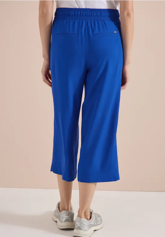 Persoon van achteren in wijde, halflange blauwe broek met elastische taille en witte sneakers.