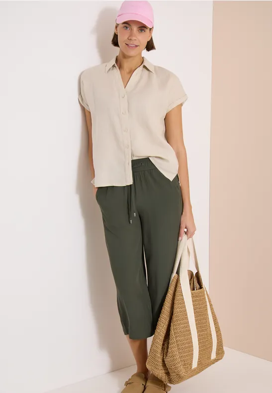 Femme en chemise blanche et pantalon vert 7/8, portant un grand sac de plage tressé, fond clair.