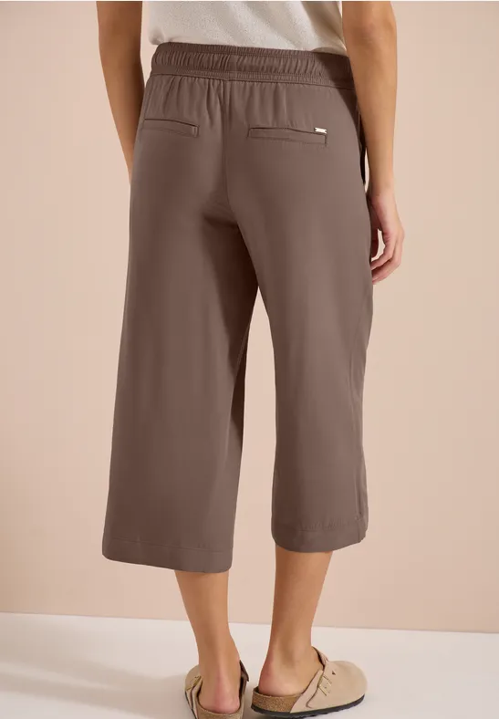 Vrouw van achter in bruine, kuitlange culottebroek met elastische taille, steekzakken en sandalen.