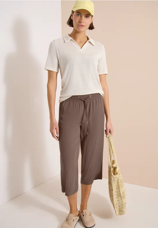 Vrouw in wit poloshirt en bruine 7/8-broek met gevlochten boodschappentas, casual look.