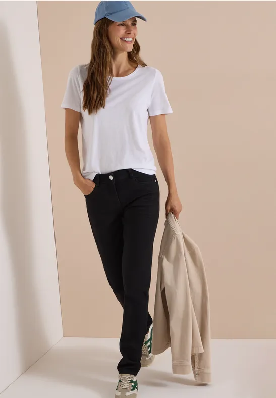 Femme en t-shirt blanc, jean noir et casquette bleue, tenant une veste beige devant un fond neutre.