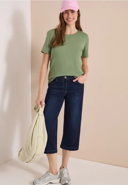 Person mit grünem T-Shirt, dunkelblauen Culotte-Jeans, weißen Sneakern und heller Tasche vor beigem Hintergrund.