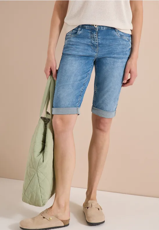 Model draagt lichtblauwe jeansbermuda, wit topje, beige loafers en een lichtgroene gewatteerde tas.