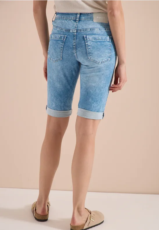 Vrouw van achteren in lichtblauwe, knielange jeansshorts met omslag en beige loafers tegen een lichte achtergrond.