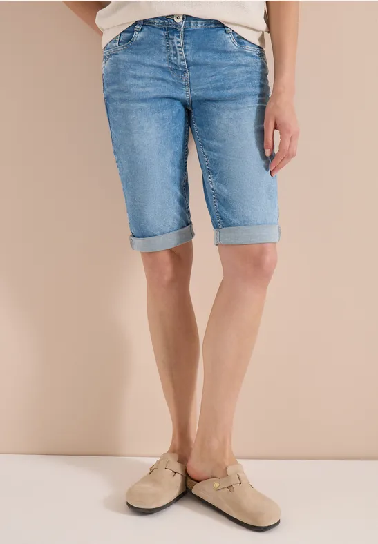 Persoon draagt lichtblauwe, omgeslagen denim bermudashort en beige instappers voor een beige achtergrond.
