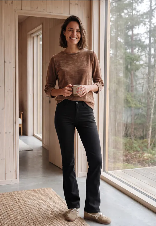 Eine Frau steht in einem Holzhaus, hält eine Tasse und lächelt.