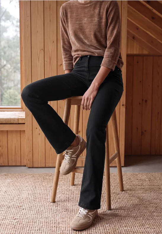 Eine Frau sitzt entspannt auf einem Holzschemel, trägt schwarze Jeans, braunen Pullover und helle Sneaker.