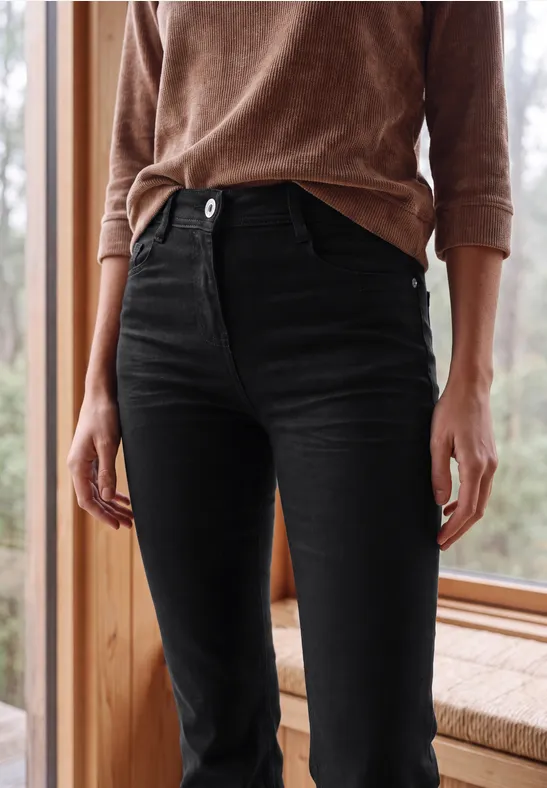 Schlanke schwarze Jeans und brauner Pullover vor einem Holzfensterrahmen.