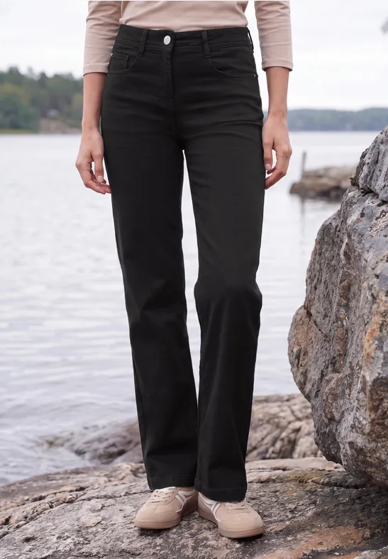 Eine Person steht am Ufer auf Felsen, trägt schwarze, gerade geschnittene Hose und beige Schuhe.