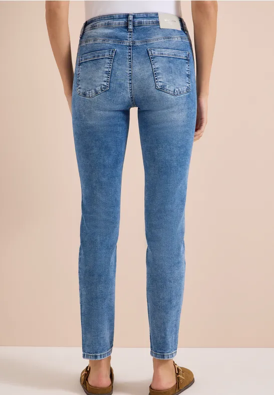 Rückansicht einer Person in hautengen blauen Jeans und Sandalen vor beigem Hintergrund.