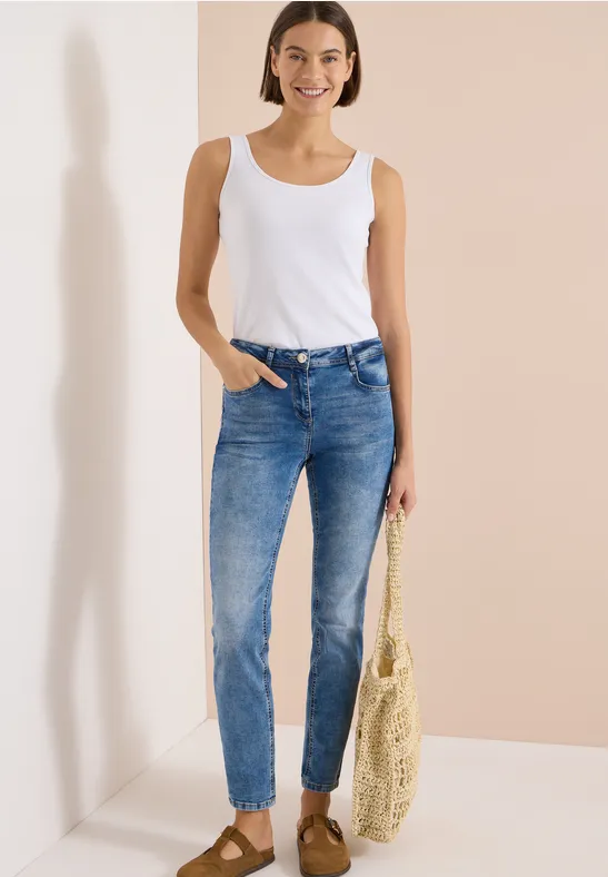 Eine Frau in blauen Jeans und weißem Tanktop mit Basttasche vor hellem Hintergrund.