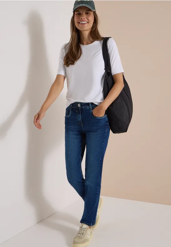 Lächelnde Frau in weißem T-Shirt, blauen Jeans, grüner Mütze und schwarzer Tasche vor heller Wand.