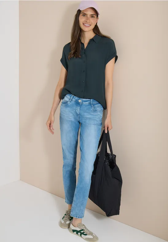 Femme en chemisier vert foncé, jean bleu, baskets vertes et sac noir sur fond beige.