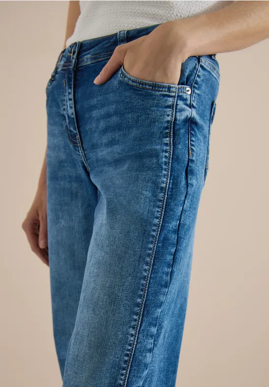 Une personne porte un jean bleu et un débardeur blanc, main dans la poche.