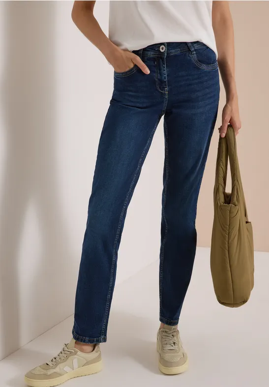 Frau trägt dunkelblaue Jeans, weißes Shirt und helle Sneaker, hält grüne Stofftasche.