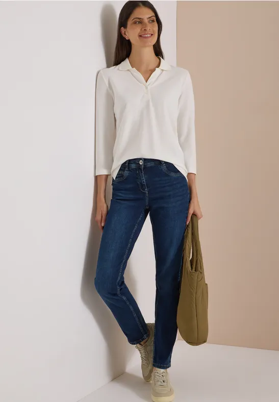 Frau lehnt lächelnd an Wand, trägt weiße Bluse, blaue Jeans und hält eine olivgrüne Tasche.