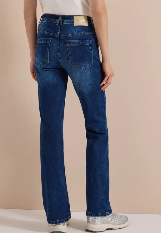 Gerade geschnittene blaue Jeans mit Label-Patch und weißen Sneakers, Rückansicht vor neutralem Hintergrund.
