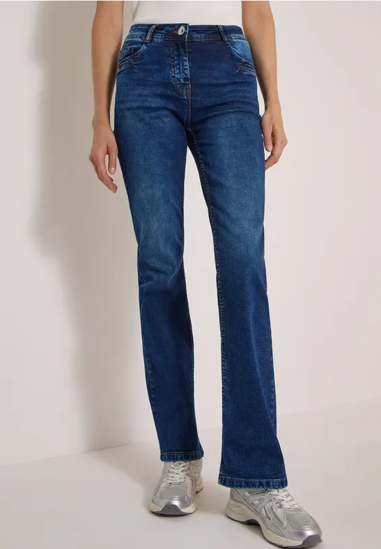 Eine Person trägt dunkelblaue gerade Jeans und graue Sneaker vor hellem Hintergrund.