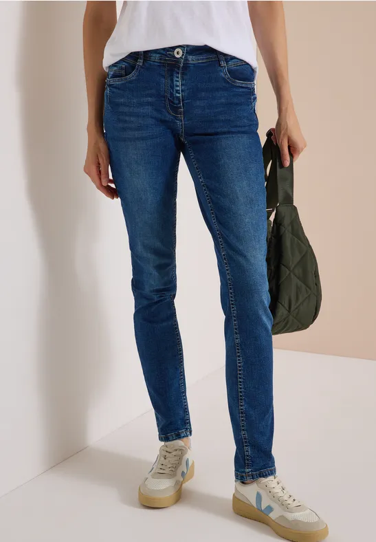 Person in enger blauer Jeans, weißen Sneakers und hält eine gesteppte grüne Tasche.