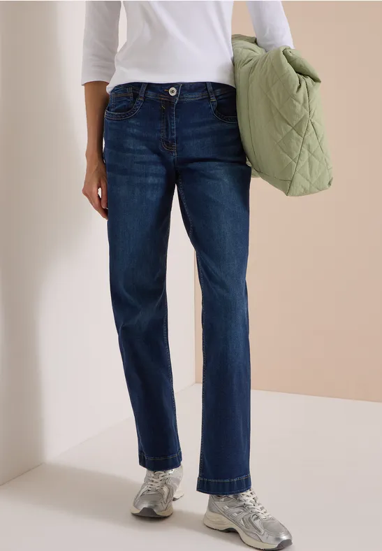 Person trägt dunkle Jeans, weißes Oberteil, silberne Sneakers und hält eine grüne gesteppte Tasche.
