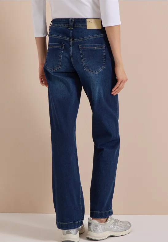 Rückenansicht einer Person in dunkelblauen, weiten Jeans und weißen Sneakers vor beigem Hintergrund.