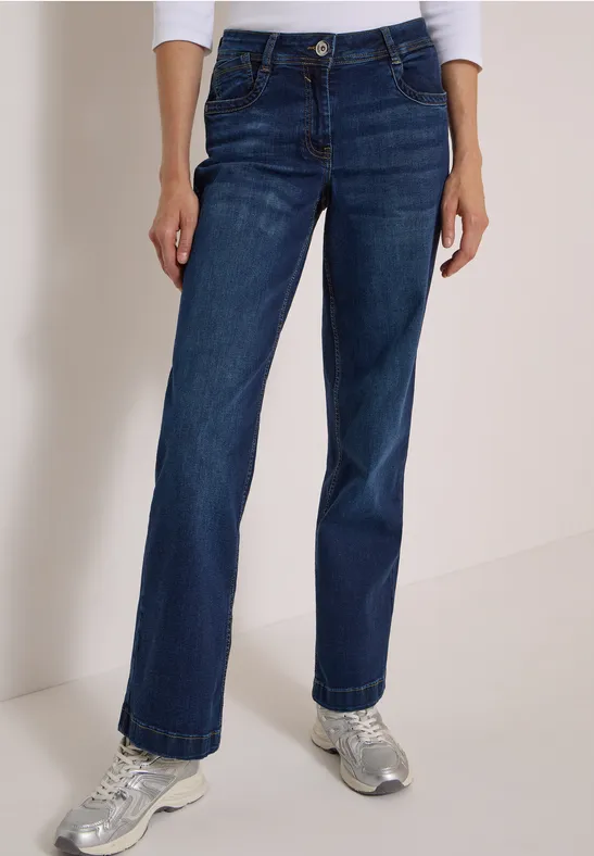 Frau trägt weite dunkelblaue Jeans und silberne Sneakers vor neutralem Hintergrund.