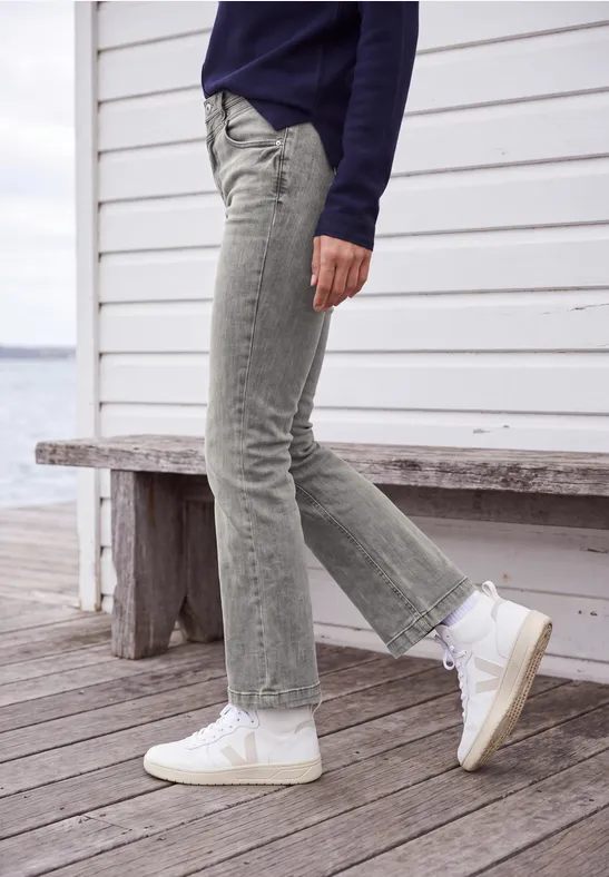 Vrouw in lichtgrijze jeans en witte sneakers op een houten dek voor een wit gebouw met uitzicht op het water.