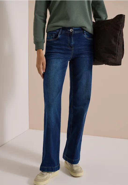Person trägt dunkelblaue, ausgestellte Jeans, grünes Langarmshirt und hält ein dunkles Kissen.