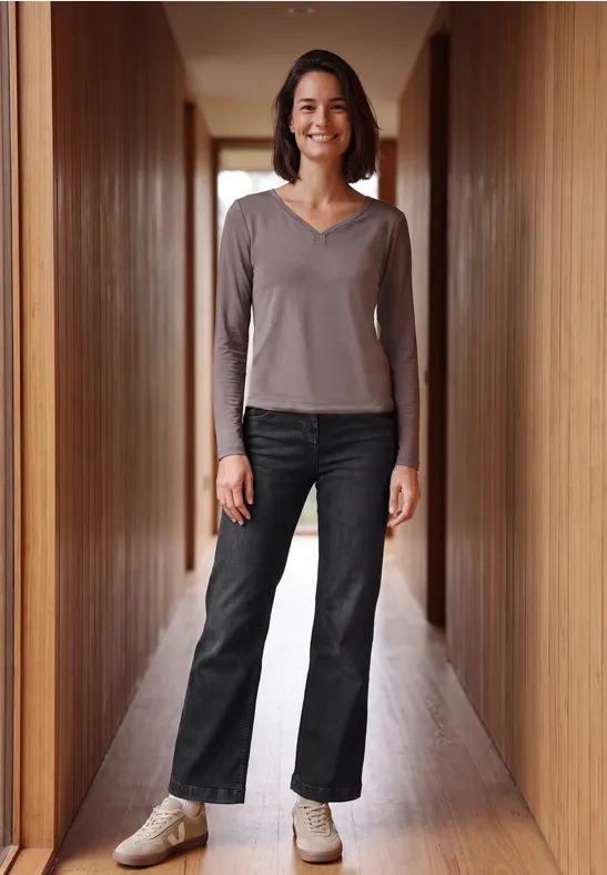 Een vrouw staat glimlachend in een houten gang, draagt een donkere jeans en een taupe longsleeve.