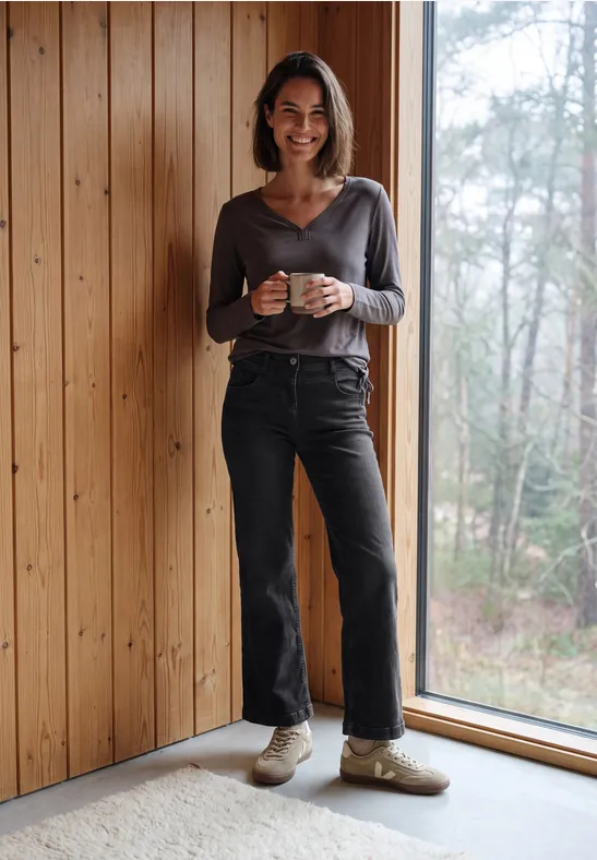 Een lachende vrouw staat in een houten huis bij het raam en houdt een mok vast.
