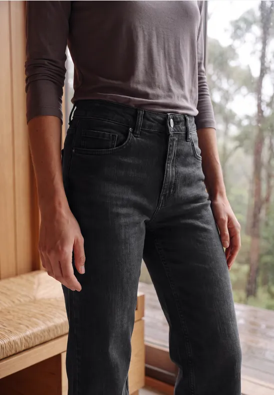 Vrouw in een grijs shirt met lange mouwen en zwarte high-waist jeans, staand voor raam met boszicht.
