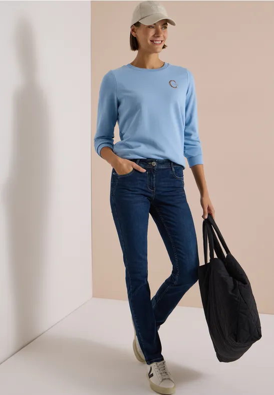 Frau mit hellblauem Shirt, dunklen Jeans, weißer Kappe und großer schwarzer Tasche lächelt entspannt.