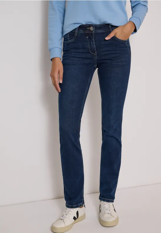 Frau in dunkelblauen Slim-Fit-Jeans, hellblauem Oberteil und weißen Sneakern vor neutraler Wand.