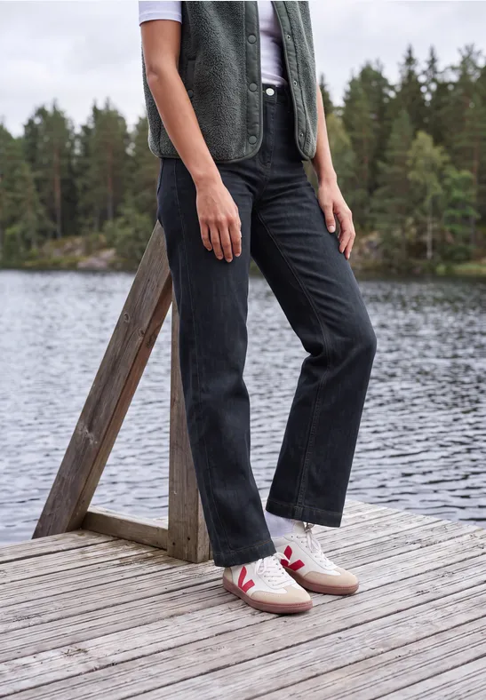 Eine Person steht auf einem Holzsteg am See, trägt schwarze Hose, weiße Sneakers mit rotem Streifen und grüne Weste.