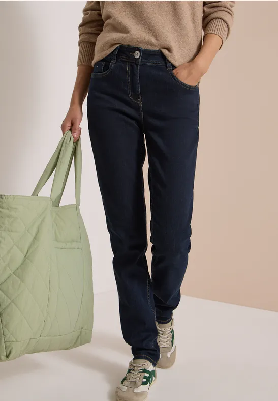 Person in dunklen Jeans, beigem Pullover und Sneakern hält eine große, hellgrüne Tasche.