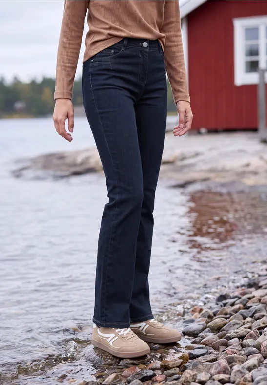 Person steht auf Kieselsteinen am Wasser, trägt dunkelblaue Jeans und beige Schuhe, im Hintergrund ein rotes Haus.