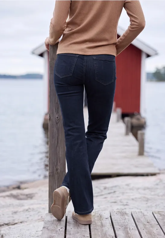Frau in Jeans und Pullover geht über Steg zum roten Bootshaus am See.