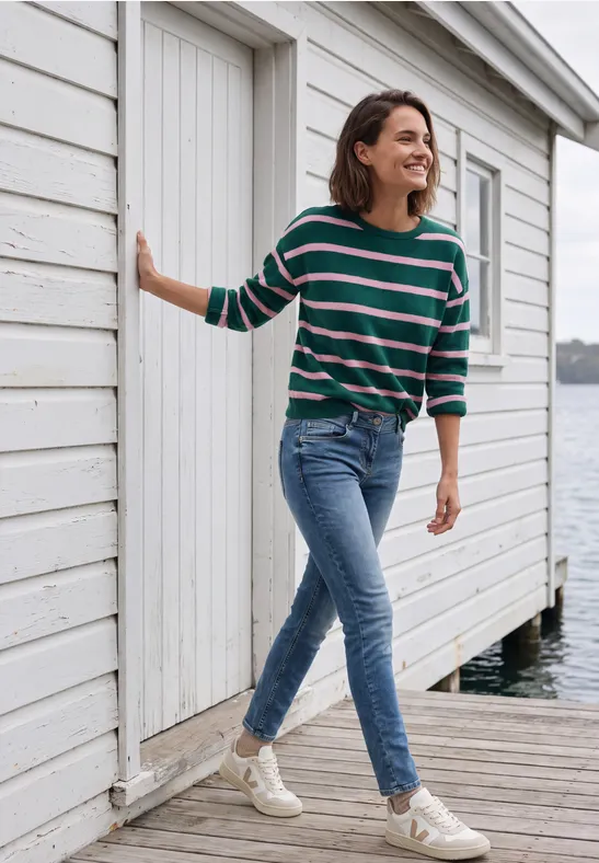 Frau in Jeans und gestreiftem Pullover spaziert lachend an einer weißen Strandhütte am Wasser entlang.