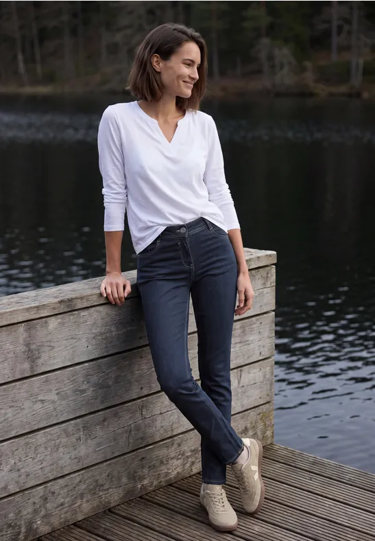 Eine Frau lehnt entspannt an einem Holzgeländer am See, trägt weiße Bluse, dunkle Hose und lächelt.