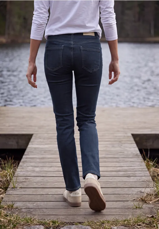 Person in engen Jeans und weißen Sneakern geht auf einem Holzsteg am See entlang.