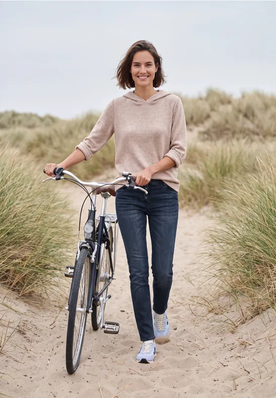Een lachende vrouw loopt met haar fiets over een zandig duinenpad, omringd door helmgras.