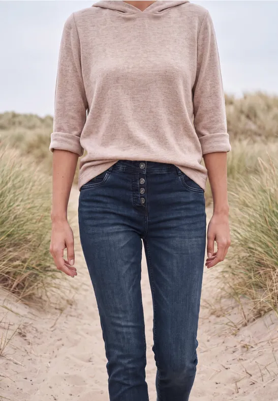 Een vrouw in een beige hoodie en blauwe jeans staand op een strand met duinen.