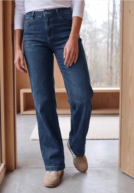 Eine Person trägt dunkle, gerade geschnittene Jeans und beigefarbene Schuhe in einem hellen Raum mit Holzwänden.
