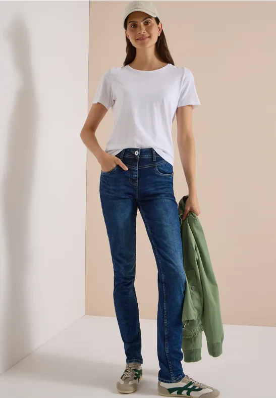 Frau in weißem T-Shirt, Jeans und Sneakern mit Kappe, hält grüne Jacke neben hellem Hintergrund.