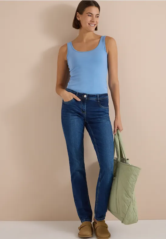 Femme en débardeur bleu et jean, portant un grand sac matelassé vert sur fond beige.