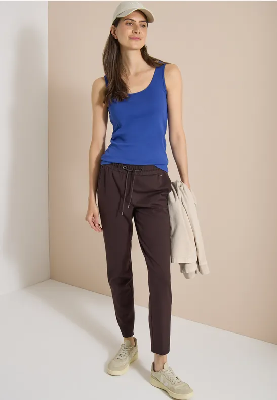 Een vrouw in een blauw tanktop, donkere broek en sneakers houdt een licht jasje vast.