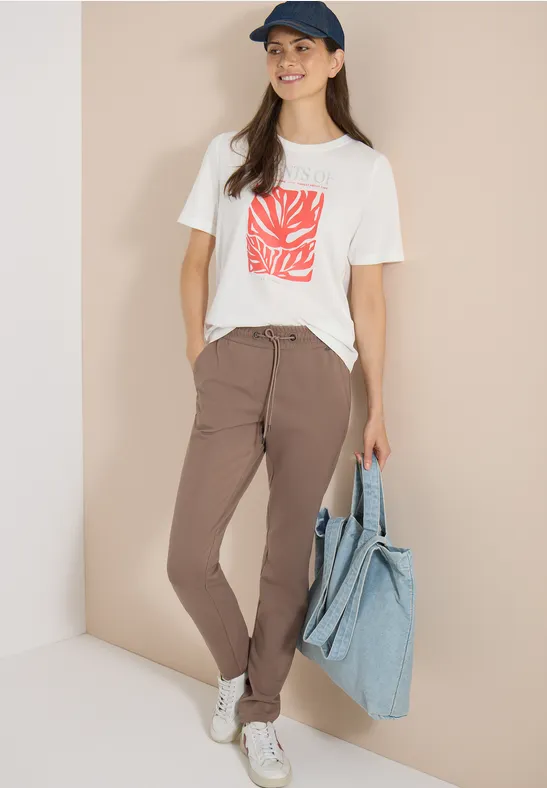 Frau trägt weiße T-Shirt mit rotem Print, braune Hose, weiße Sneaker und große blaue Tragetasche.