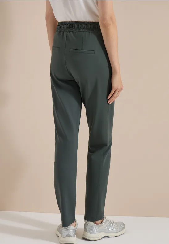 Model van achteren in een donkergroene, rechte broek met elastische taille en sneakers.